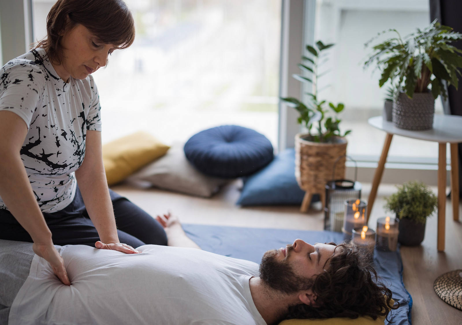 Ein Mann liegt mit dem Rücken auf einer Matte am Boden und eine Frau sitzt daneben. Sie therapiert mit Drucktechniken den Bauchbereich des Mannes.