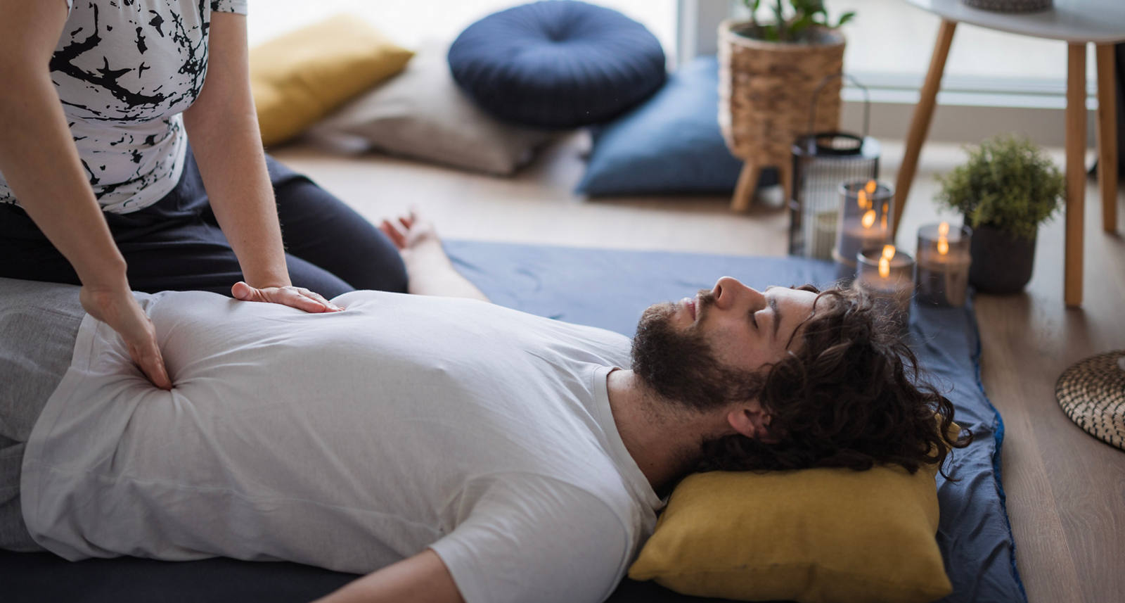 Ein Mann liegt mit dem Rücken auf einer Matte am Boden und eine Frau sitzt daneben. Sie therapiert mit Drucktechniken den Bauchbereich des Mannes.