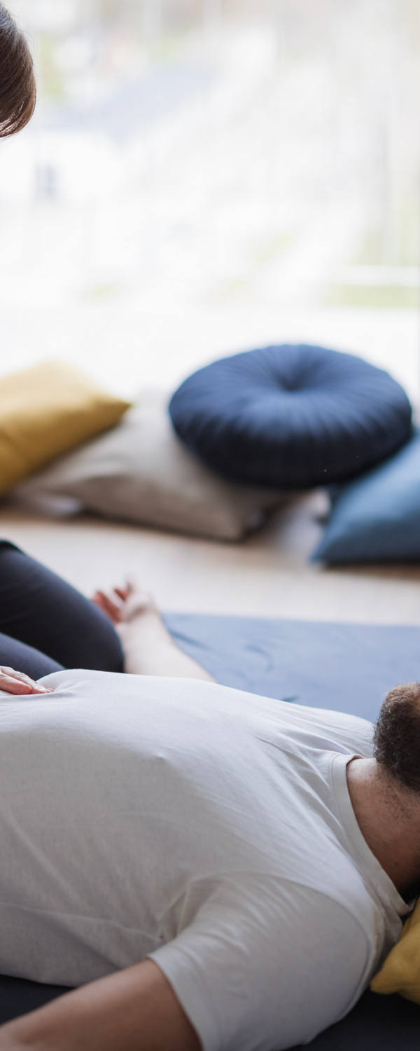 Ein Mann liegt mit dem Rücken auf einer Matte am Boden und eine Frau sitzt daneben. Sie therapiert mit Drucktechniken den Bauchbereich des Mannes.