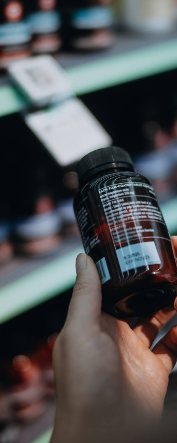 Eine Frau steht in der Drogerie vor einem Regal und hält eine Dose Medikamente in Händen während Sie die Inhaltsstoffe liest.