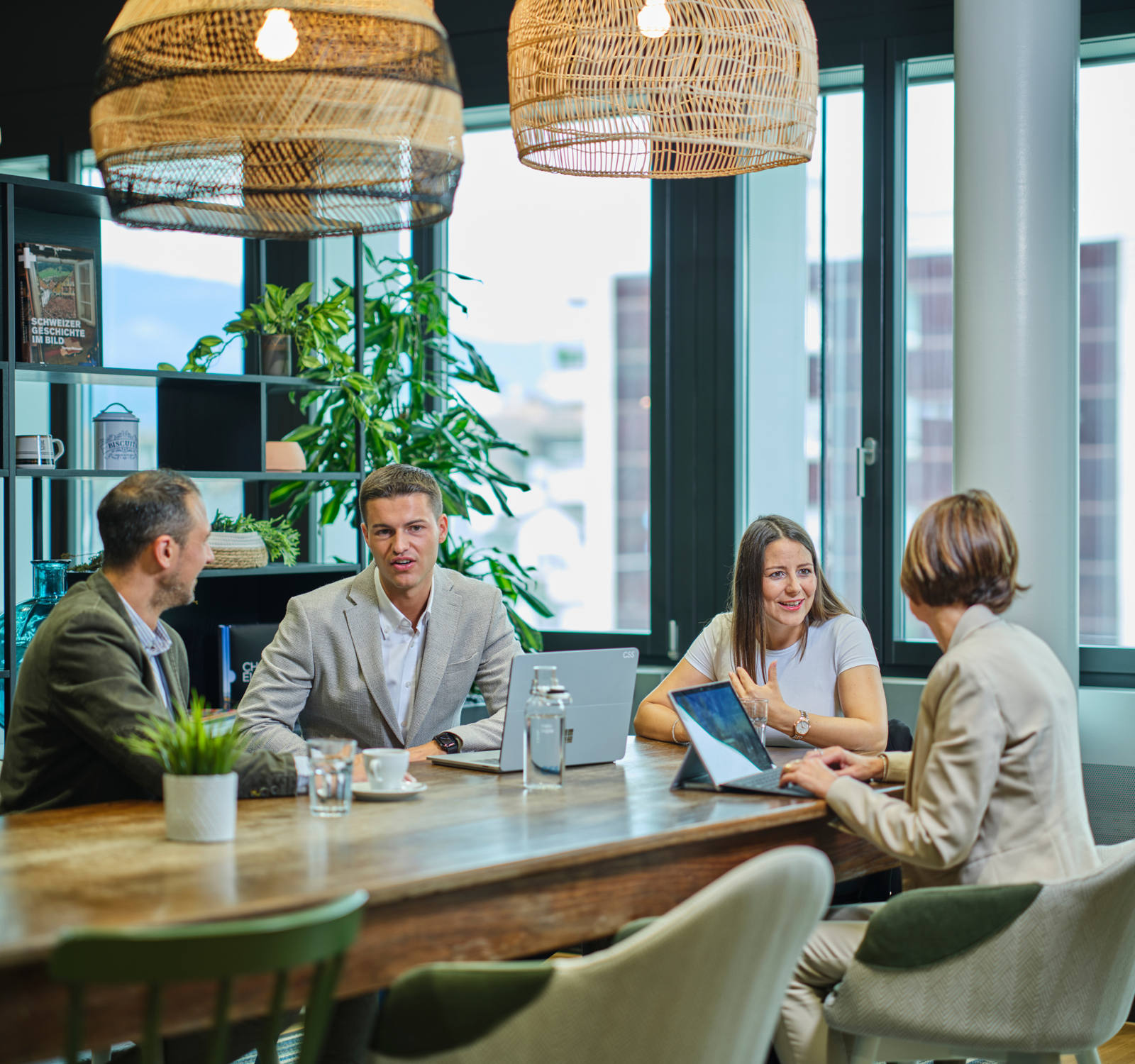 Deux femmes et deux hommes sont assis autour d’une grande table en bois et discutent de différents sujets. Deux ordinateurs portables sont posés sur la table.