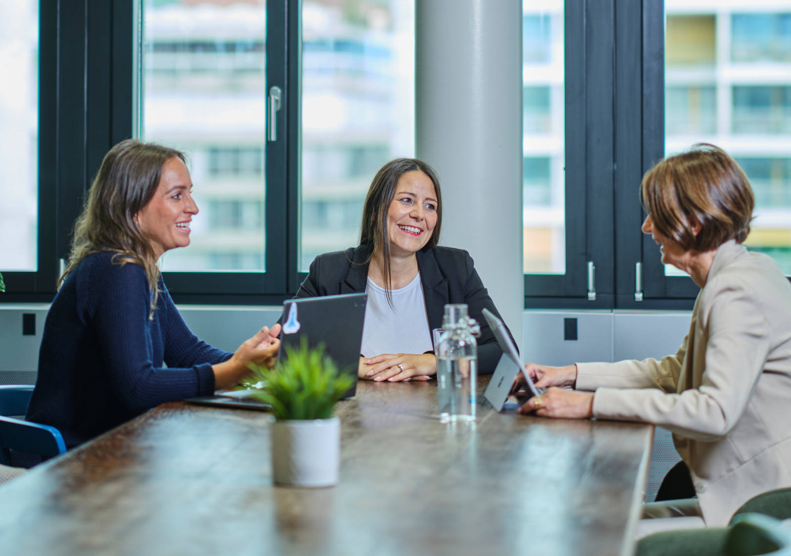 Tre donne sono sedute con i loro laptop a un grande tavolo di legno.