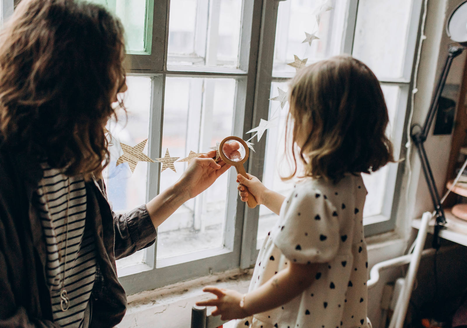 Eine Frau spielt mit ihrer Tochter am Fenster und schauen durch eine Lupe die Sternen-Girlande an.