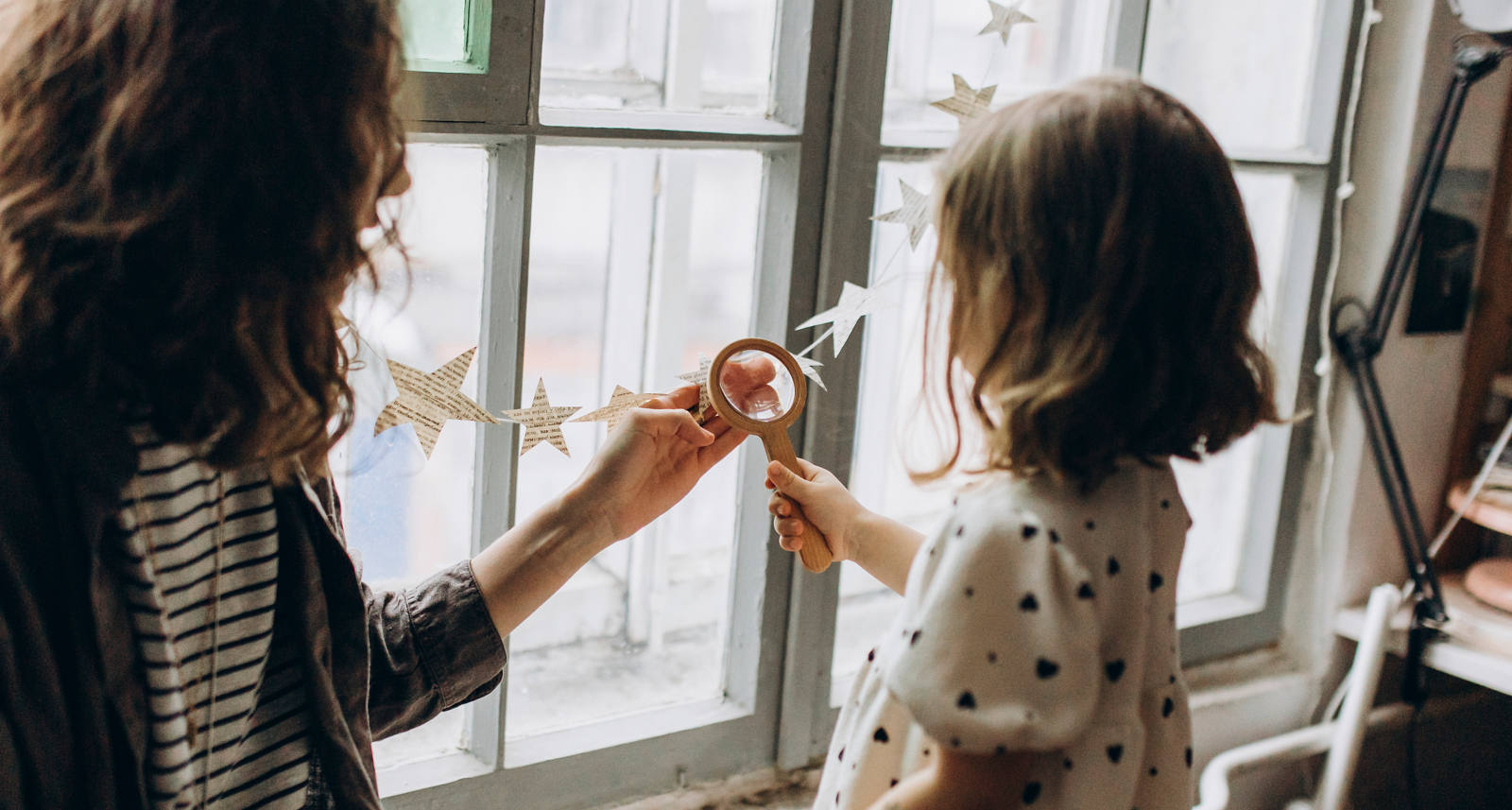 Eine Frau spielt mit ihrer Tochter am Fenster und schauen durch eine Lupe die Sternen-Girlande an.