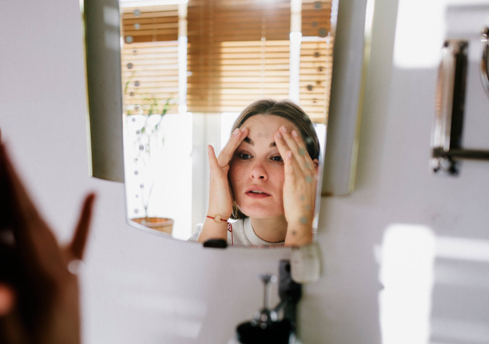 A young woman stands in the bathroom in front of a mirror, holding her hands to her temples.