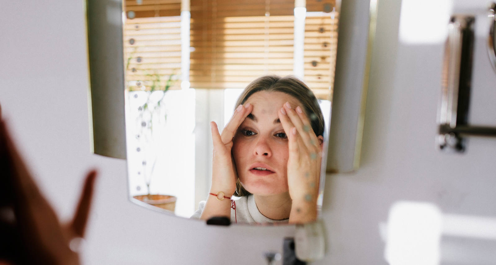 A young woman stands in the bathroom in front of a mirror, holding her hands to her temples.