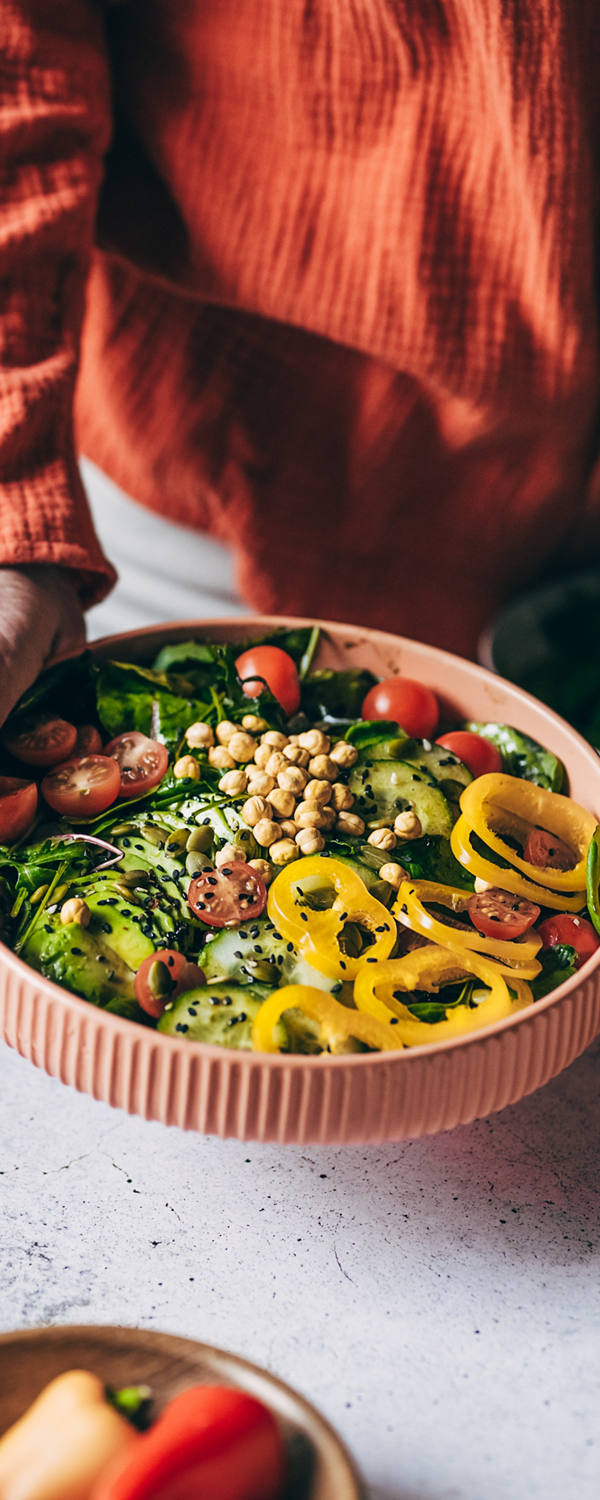 Eine Frau steht in der Küche und hält eine Salat-Bowl in die Kamera. Die Schüssel ist gefüllt mit verschiedenem und butem Gemüse und Kichererbsen.