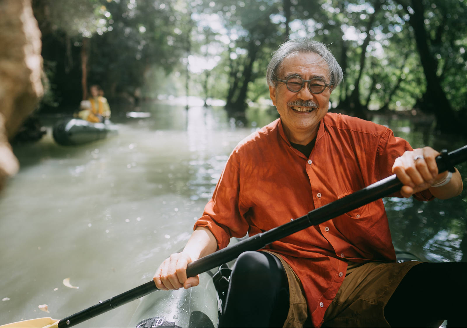An older gentleman happily sits in a kayak and paddles along a river in the sunshine along trees and rock faces.