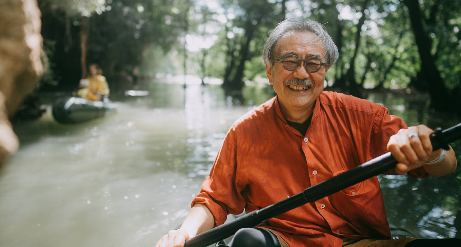Un homme âgé est joyeusement assis dans un kayak et pagaie sur une rivière le long des arbres et des parois rocheuses sous le soleil.