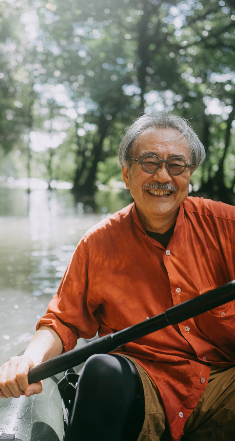 An older gentleman happily sits in a kayak and paddles along a river in the sunshine along trees and rock faces.