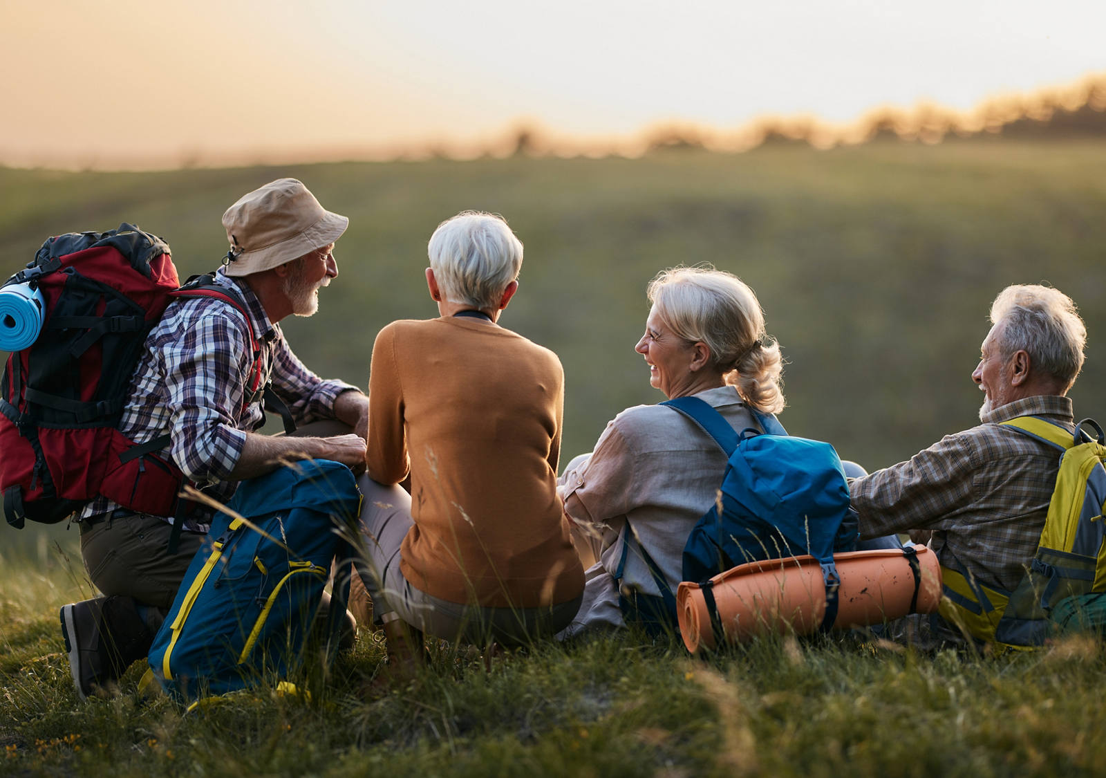 Zwei ältere Frauen und Männer mit Rucksäcken sind auf einer Wanderung und machen Pause, sitzen auf der Wiese und lachen.