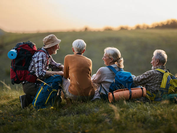 Zwei ältere Frauen und Männer mit Rucksäcken sind auf einer Wanderung und machen Pause, sitzen auf der Wiese und lachen.