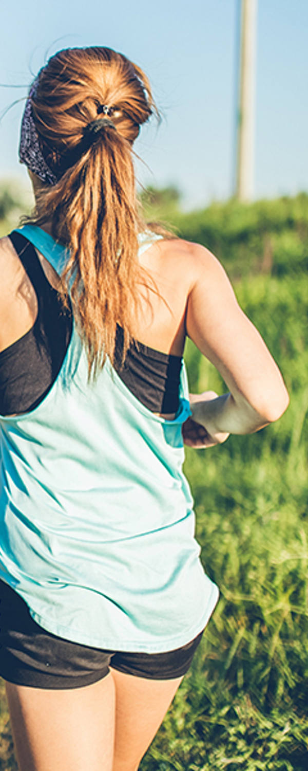 Eine junge Frau in kurzer Sportkleidung joggt in der Sonne einen Feldweg entlang.