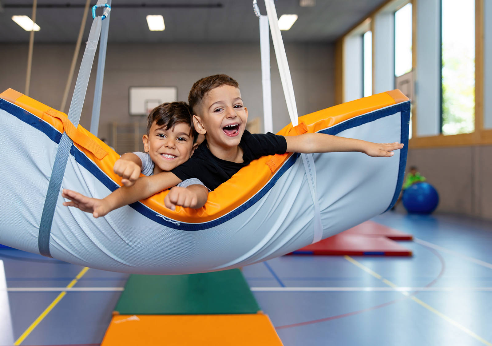 Zwei fröhliche Kinder im Primarschulalter spielen in einer Turnhalle und schaukeln auf einer hängenden Matte.