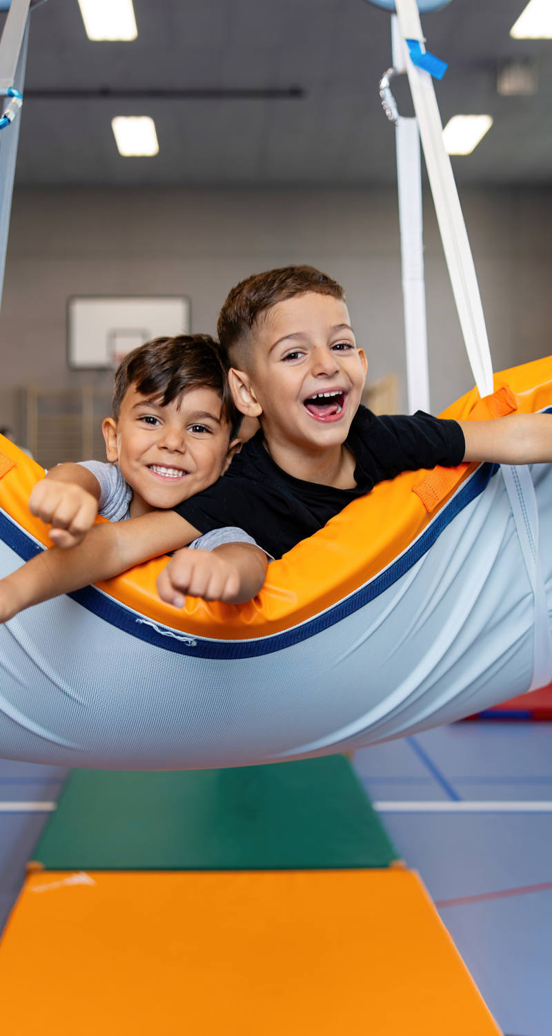 Zwei fröhliche Kinder im Primarschulalter spielen in einer Turnhalle und schaukeln auf einer hängenden Matte.