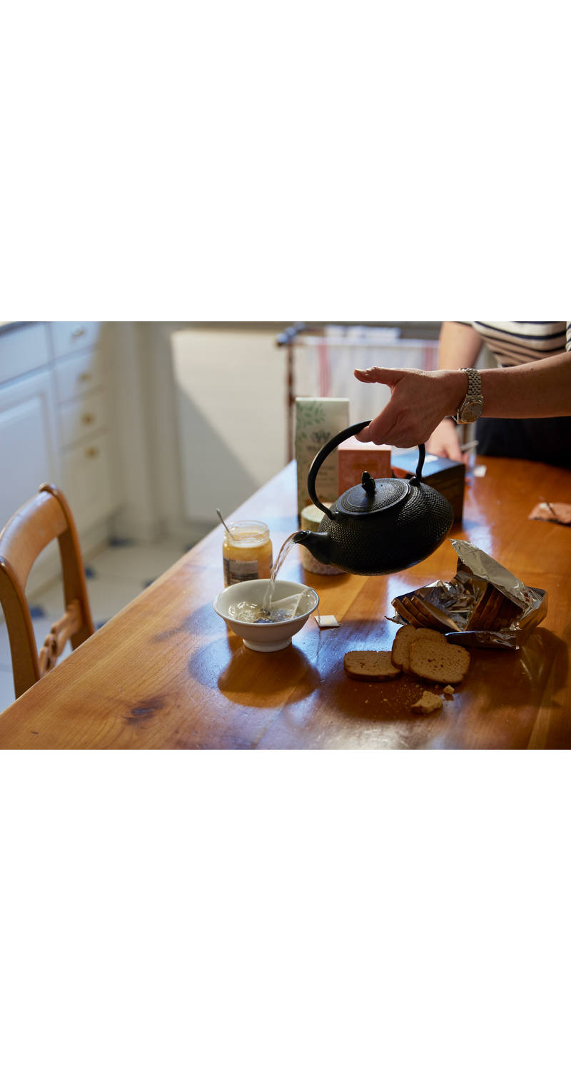 A woman pours hot water into a teacup at the kitchen table.
